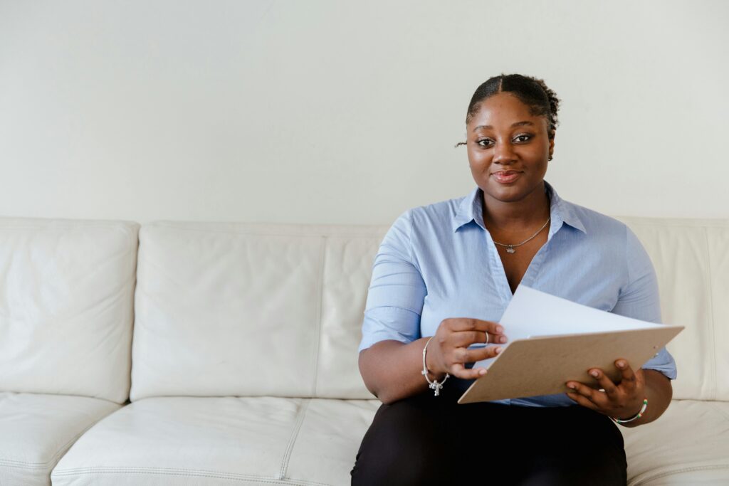 A confident woman in blue shirt sitting on a white sofa holding a clipboard, smiling.
